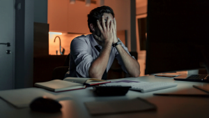 Stressed businessman sitting at his desk late at night with his hands covering his face, surrounded by paperwork, computer, and dim lighting.