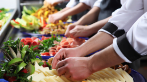 Chefs in uniform arranging an elaborate buffet with sliced cheese, meats, fruits, and fresh herbs, preparing for a professional catering event.