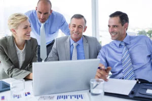 Smiling business professionals in formal attire collaborating around a laptop in a modern office with charts and reports on the table.