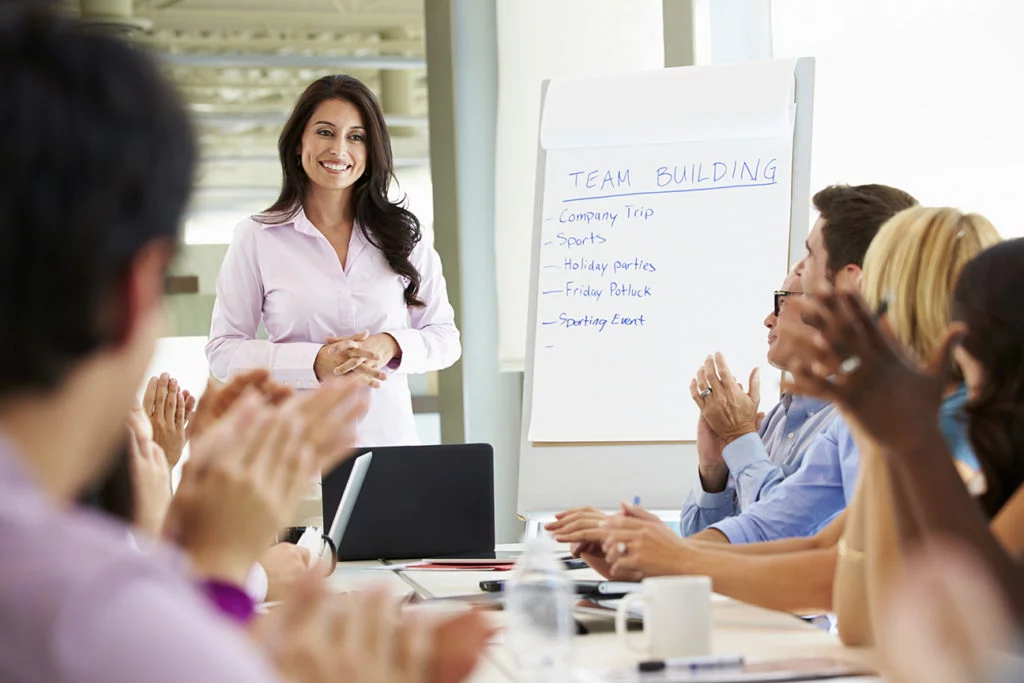 Smiling female presenter leading a team building workshop with a flipchart in a modern office, while coworkers applaud and engage in the session.