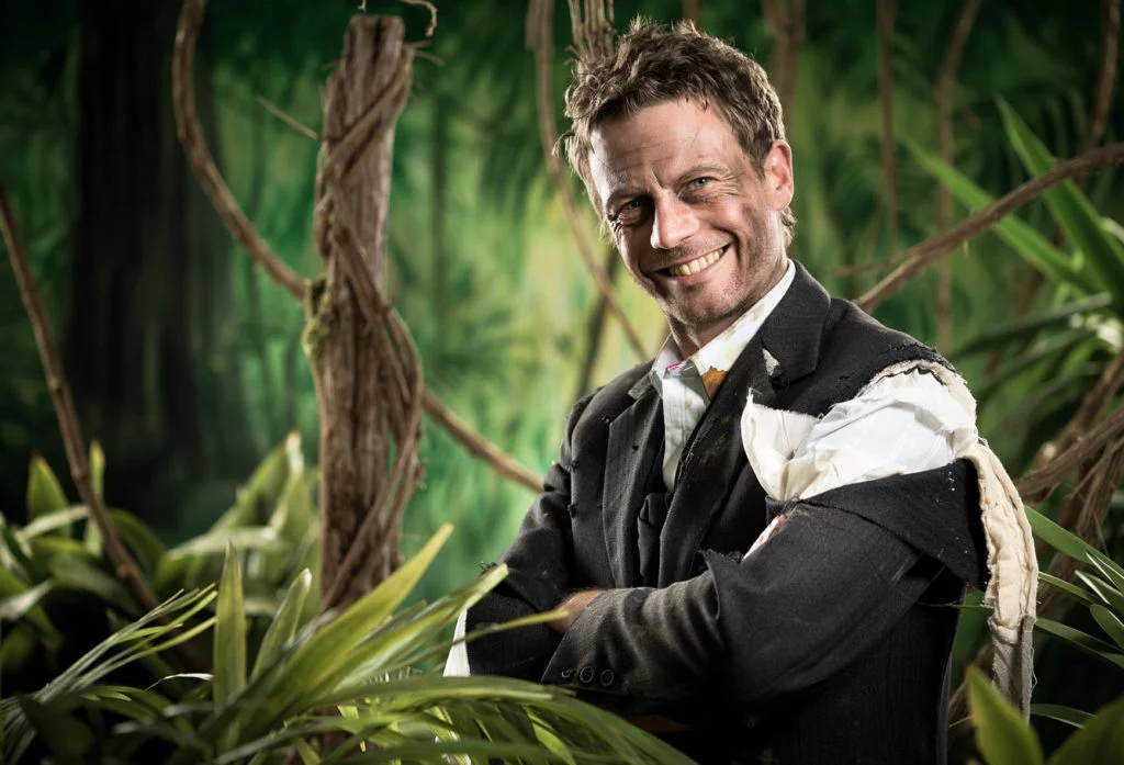 Smiling man in a torn business suit standing in a jungle, symbolizing survival and resilience in challenging environments