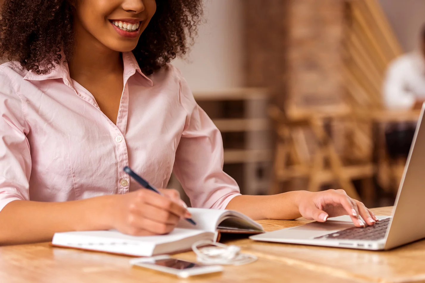 Smiling woman in a pink shirt studying with a notebook and laptop at a wooden table, multitasking between writing and typing in a cozy indoor setting.