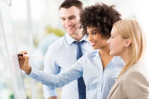 Three smiling coworkers brainstorming and writing on a whiteboard during a team meeting in a bright office