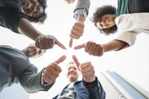 Group of smiling professionals standing in a circle giving thumbs up toward the camera, viewed from below.