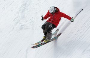 Skier in red jacket performing jump on snowy slope with colorful skis and helmet, showcasing winter sports action