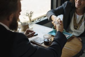 Two people shaking hands across a small table with coffee cups, indicating a business agreement