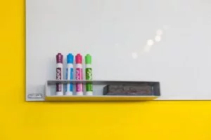 Colorful dry-erase markers and eraser on whiteboard tray with bright yellow wall