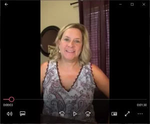 Smiling woman in patterned sleeveless top sitting indoors in front of purple curtains, paused video screenshot