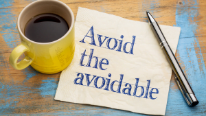 Yellow coffee mug filled with black coffee next to a napkin with the text "Avoid the avoidable" and a silver pen on a rustic wooden table