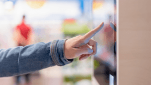 Person in denim jacket using touchscreen kiosk with finger pointing at screen in public space