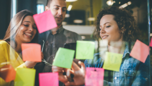 A group of diverse young professionals smiling and collaborating while sticking colorful sticky notes on a glass wall during a brainstorming session