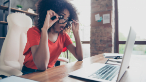 Young woman with curly hair and glasses looking shocked at laptop screen in home office