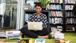 Businesswoman working on laptop sitting on floor in front of bookshelf