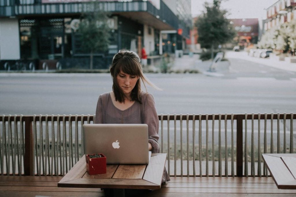Young woman focused working on a laptop outdoors at wooden table near a city street
