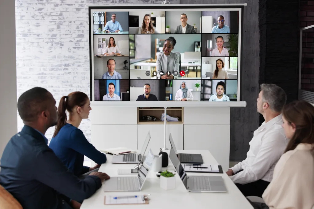 Business team sitting in conference room watching a large screen with a virtual meeting of diverse professionals