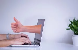 Laptop with typing hands and a 3D thumbs-up hand emerging from the screen, white desk and green plant
