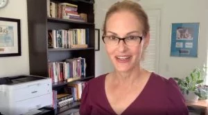 Woman wearing glasses speaking on video from home office with bookshelf and printer in background