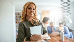 Smiling woman holding a tablet in an office setting with colleagues working in the background, depicting a confident professional or trainer