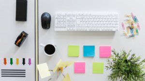 White wireless keyboard, black mouse, smartphone, colorful paper clips, sticky notes and a cup of black coffee on a white desk workspace