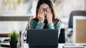Woman in glasses rubbing her eyes while sitting at a desk with a laptop, showing signs of fatigue or eye strain