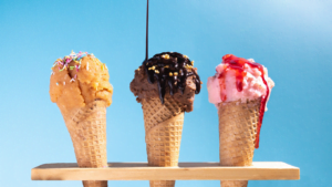 Three ice cream cones with colorful scoops topped with sprinkles, chocolate sauce, and strawberry syrup against a bright blue background