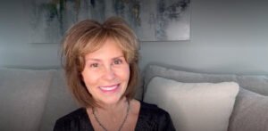 Middle-aged woman sitting indoors on a gray couch with cushions, smiling warmly at the camera