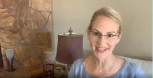 Woman wearing glasses speaking in a warmly decorated living room with stone wall and lamp