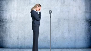 Businesswoman in dark suit standing with head in hand in front of a microphone against a gray wall, expressing stress or anxiety