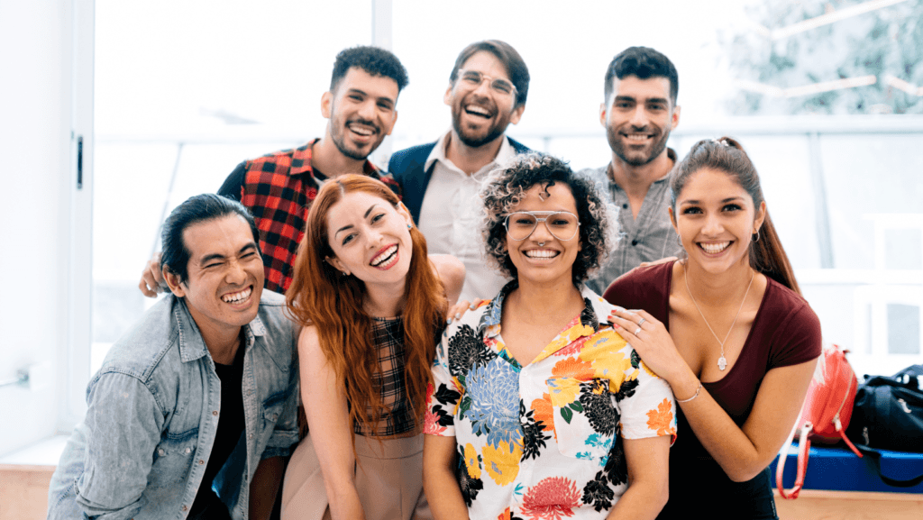 A diverse group of smiling young adults posing together indoors, showing a sense of friendship, teamwork, and positive energy in a modern setting.