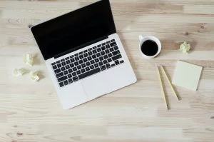 Overhead view of a laptop on a wooden desk with coffee cup, two pencils, notepad, and crumpled paper balls