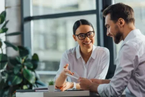 Businesswoman wearing glasses smiling and discussing documents with male colleague in bright modern office