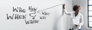 Woman writing on a whiteboard with question words who, how, what, when, where, and why during a business presentation