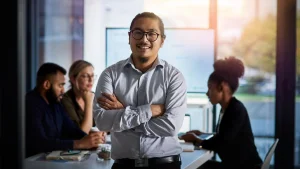 Confident young Asian businessman standing with arms crossed in a modern office, with colleagues working in the background