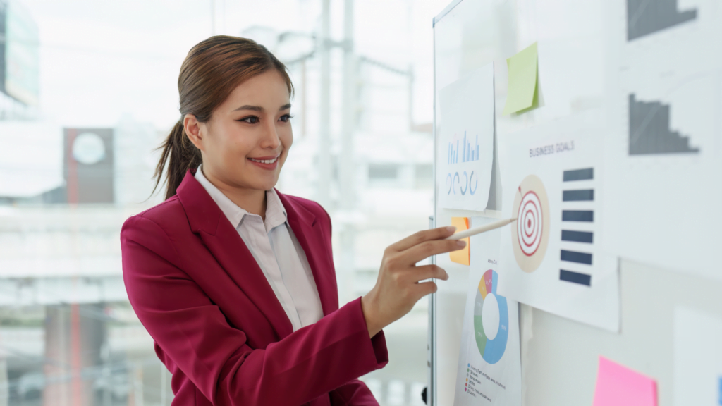 Smiling businesswoman in red blazer pointing at business goals charts on a whiteboard during a presentation