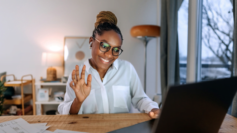 Woman waving and smiling during a video call in a cozy home office environment