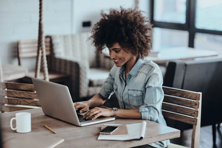 Smiling woman working on laptop at home with coffee, phone, and notebook on the table