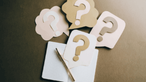 Notepad with question mark speech bubbles and silver pen on wooden desk