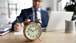 Focused businessman in suit writing notes with vintage alarm clock in foreground on office desk