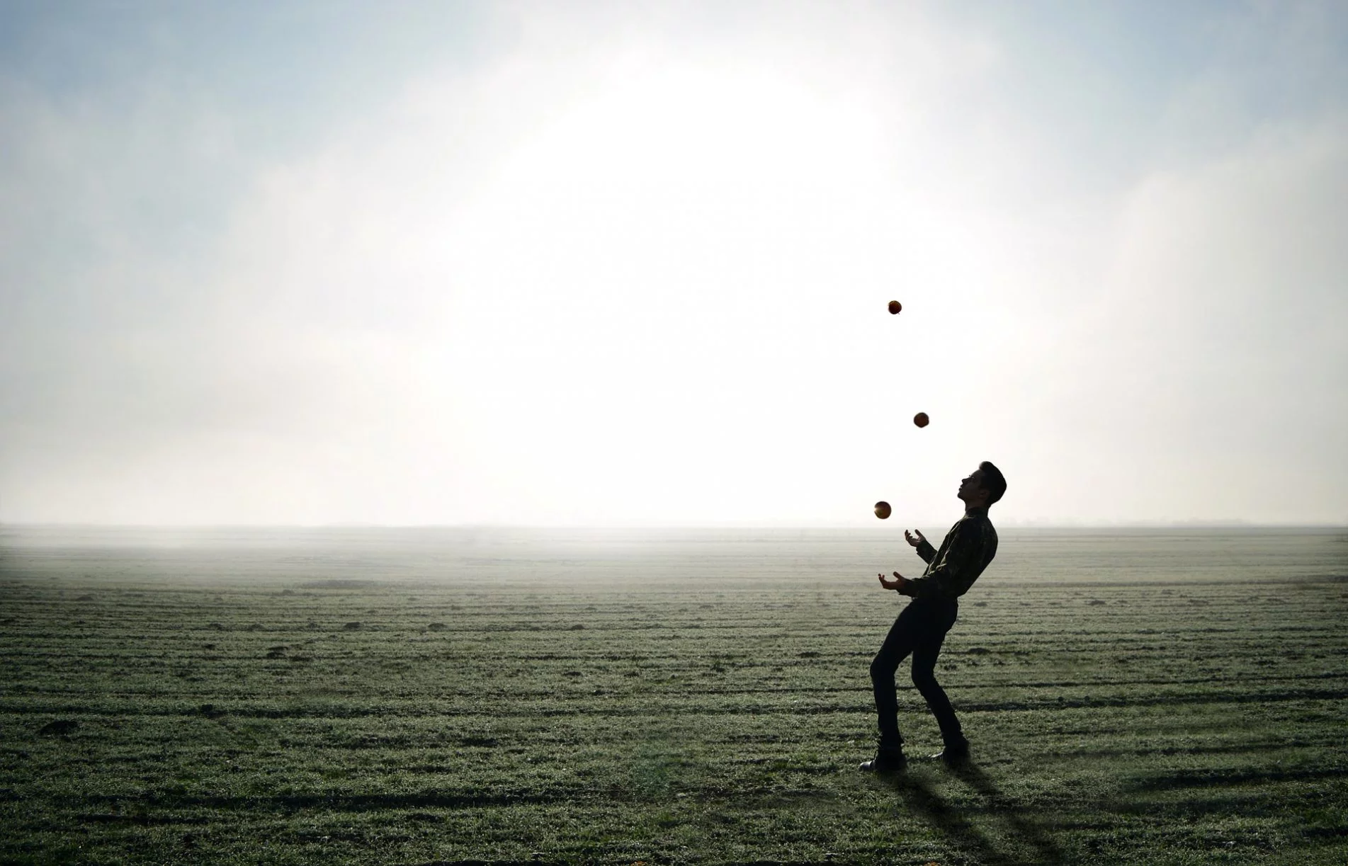 Man juggling three balls outdoors on a foggy field with bright sky in the background