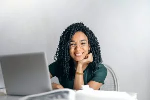 A smiling woman with dreadlocks sits at a desk, working on a laptop