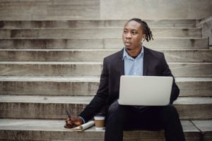 Businessman sitting on outdoor steps with laptop, writing notes and thinking about work